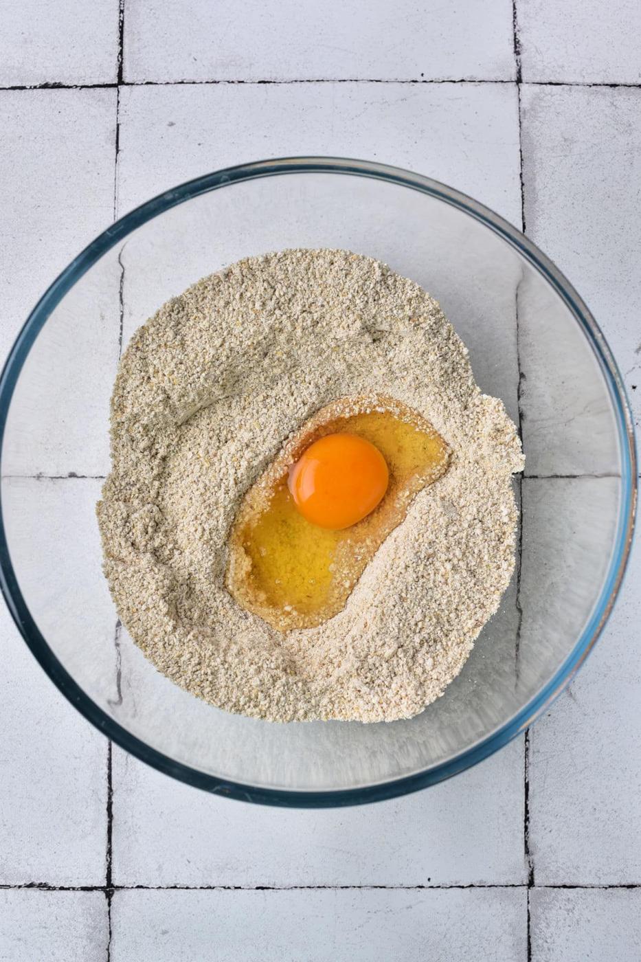 A glass bowl with matzo and an egg in the center sits on a tiled surface, reminiscent of the beginnings of a matzo ball preparation.