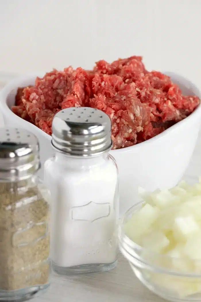 Bowl of ground beef, diced onions in a small bowl, and salt and pepper shakers on a light surface—perfect inspiration if you're wondering what to cook with ground beef.