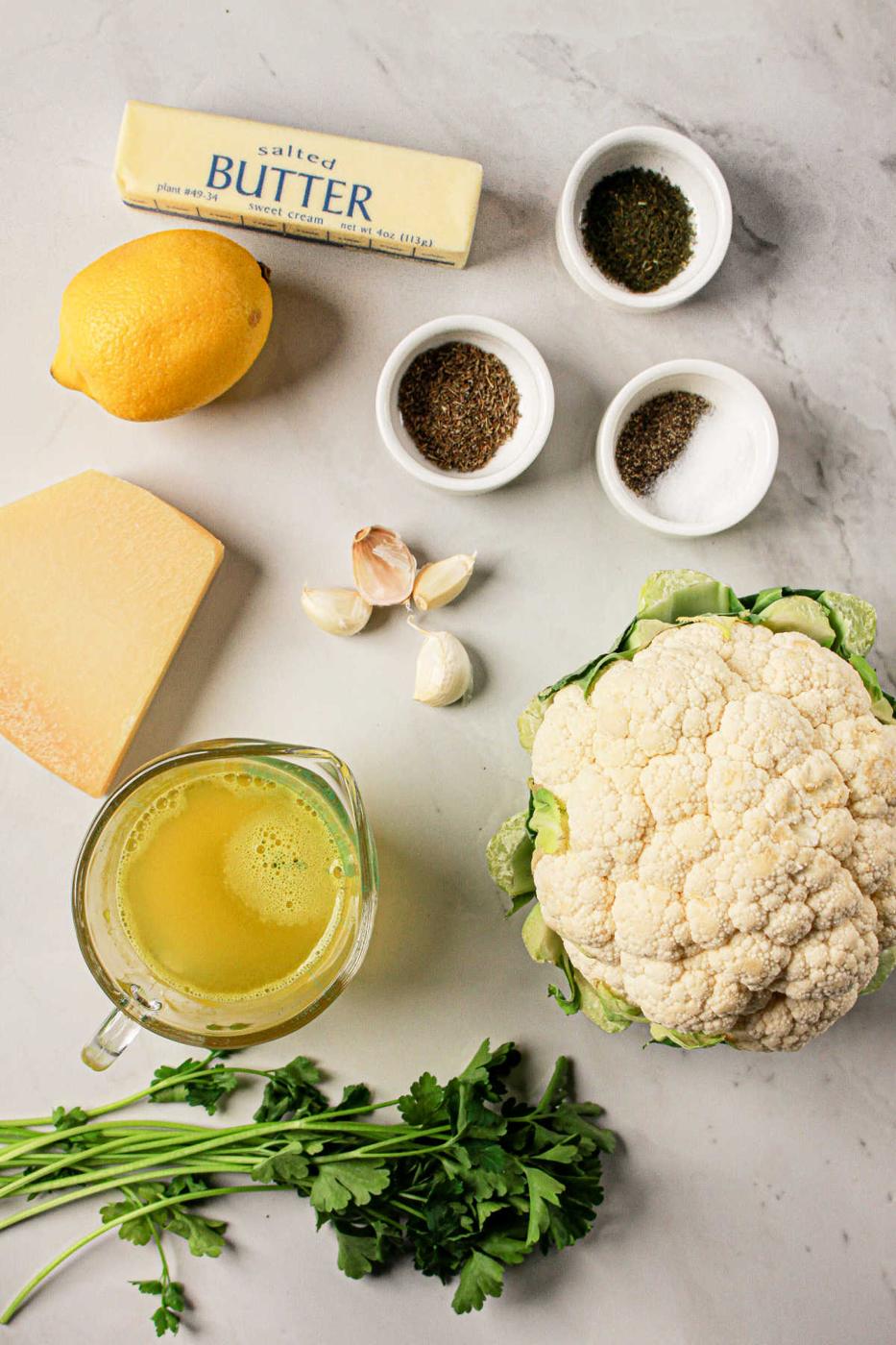Ingredients on a marble surface: butter, lemon, cheese, garlic, whole cauliflower, herbs, broth in a jug, and small spice bowls. Perfect for creating a delightful cauliflower recipe that can easily be adapted for Crock Pot cooking.