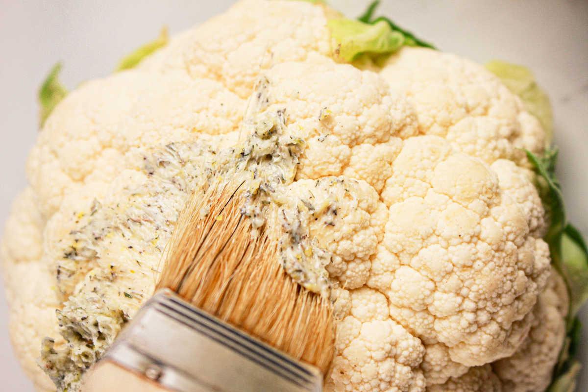 Close-up of a whole cauliflower being brushed with seasoned oil or butter using a flat brush, perfect for a crock pot transformation.