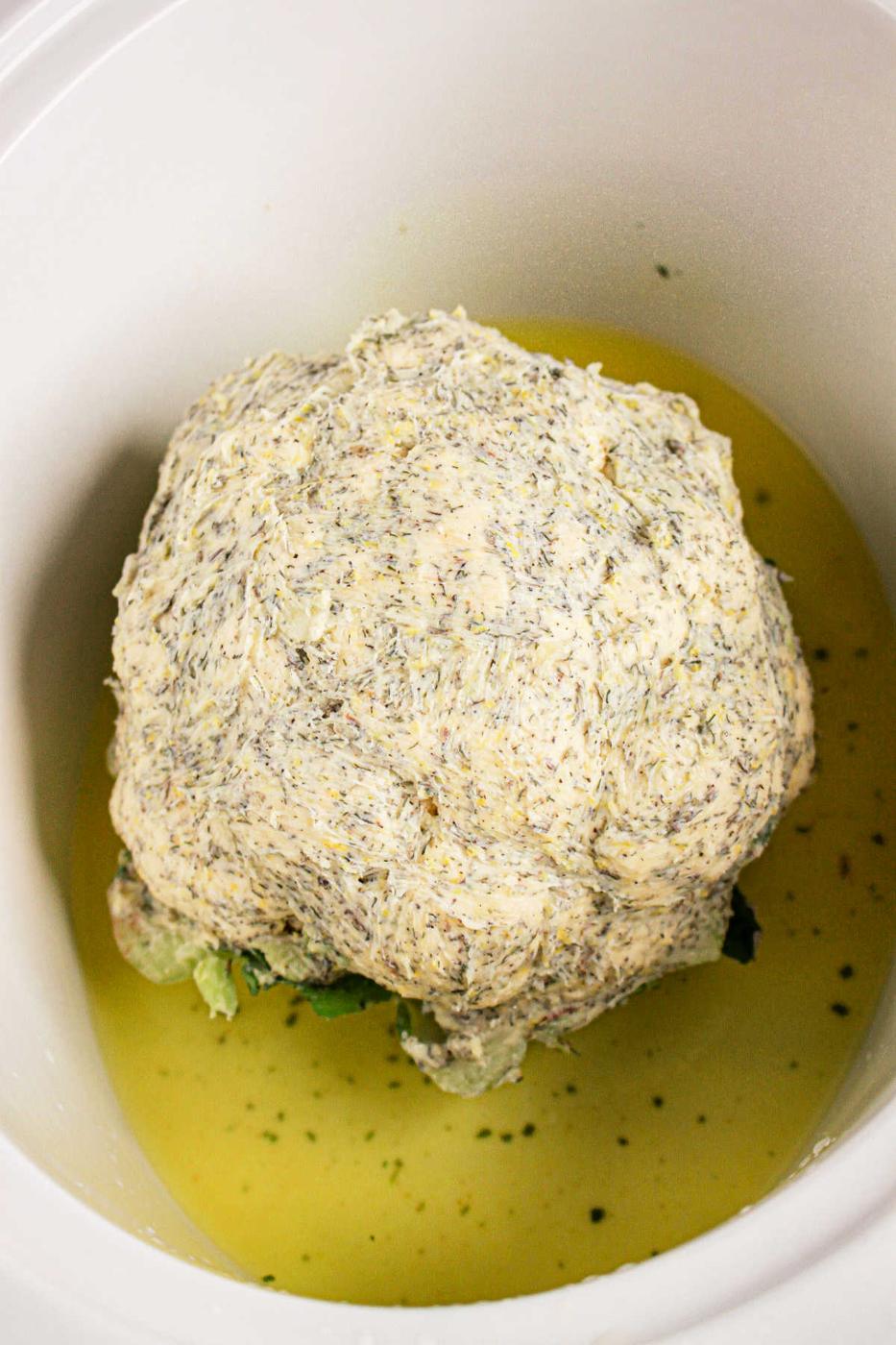 A whole cauliflower head rests in a crock pot, gently coated with a seasoned sauce and partially submerged in flavorful liquid.
