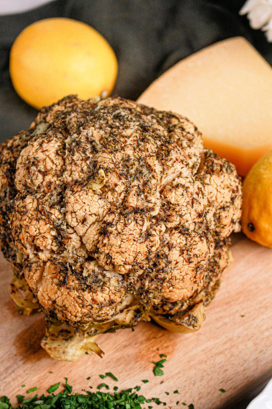 A whole roasted cauliflower, seasoned with herbs, sits on a wooden board surrounded by lemons and cheese. This delightful recipe showcases the versatility of this humble vegetable, perfect for savoring as is or prepared in a crock pot for tender perfection.