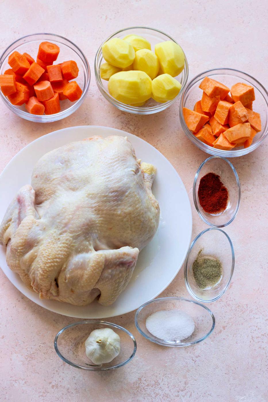 Whole Chicken Brine, carrots, potatoes, sweet potatoes, garlic, and spices are arranged in bowls on a light surface.