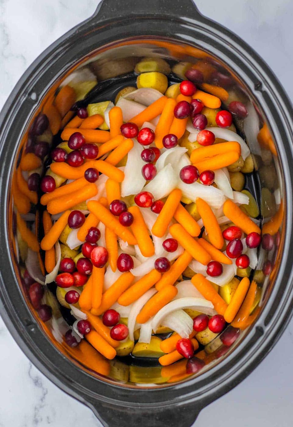 A black slow cooker filled with a whole chicken, carrots, cranberries, sliced onions, and potatoes, all arranged in a colorful, layered mixture.