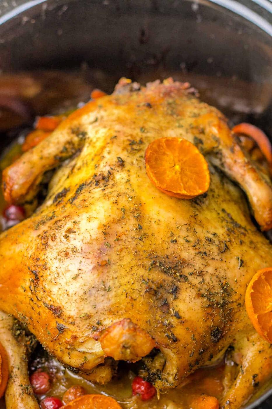 Whole roasted chicken in a pot, seasoned with herbs, garnished with orange slices, red berries, and tender potatoes, with crispy golden-brown skin.