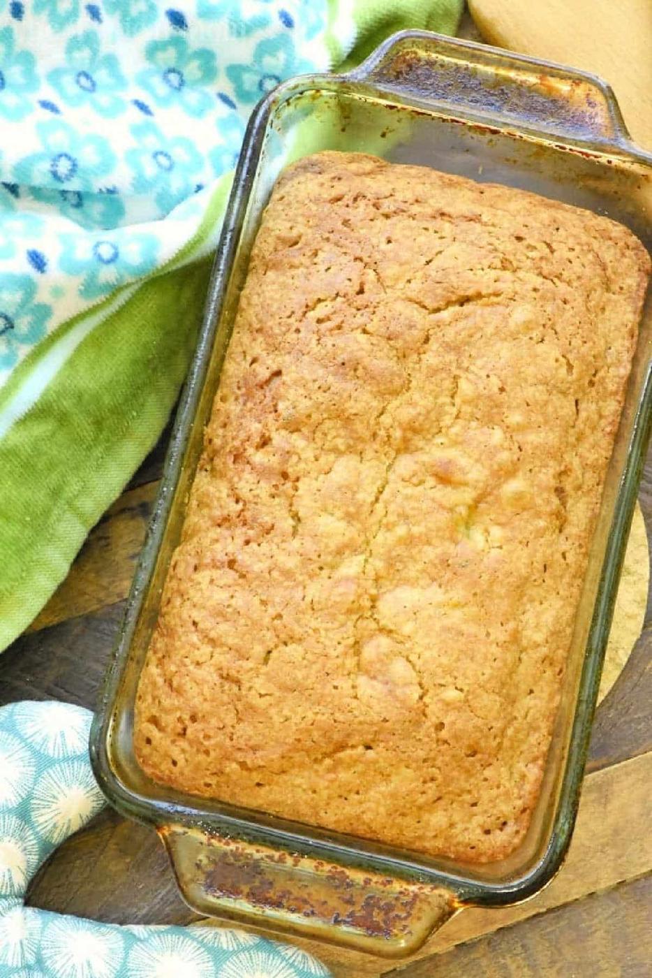 A loaf of baked zucchini pineapple bread in a glass pan rests on a wooden surface with patterned towels nearby.