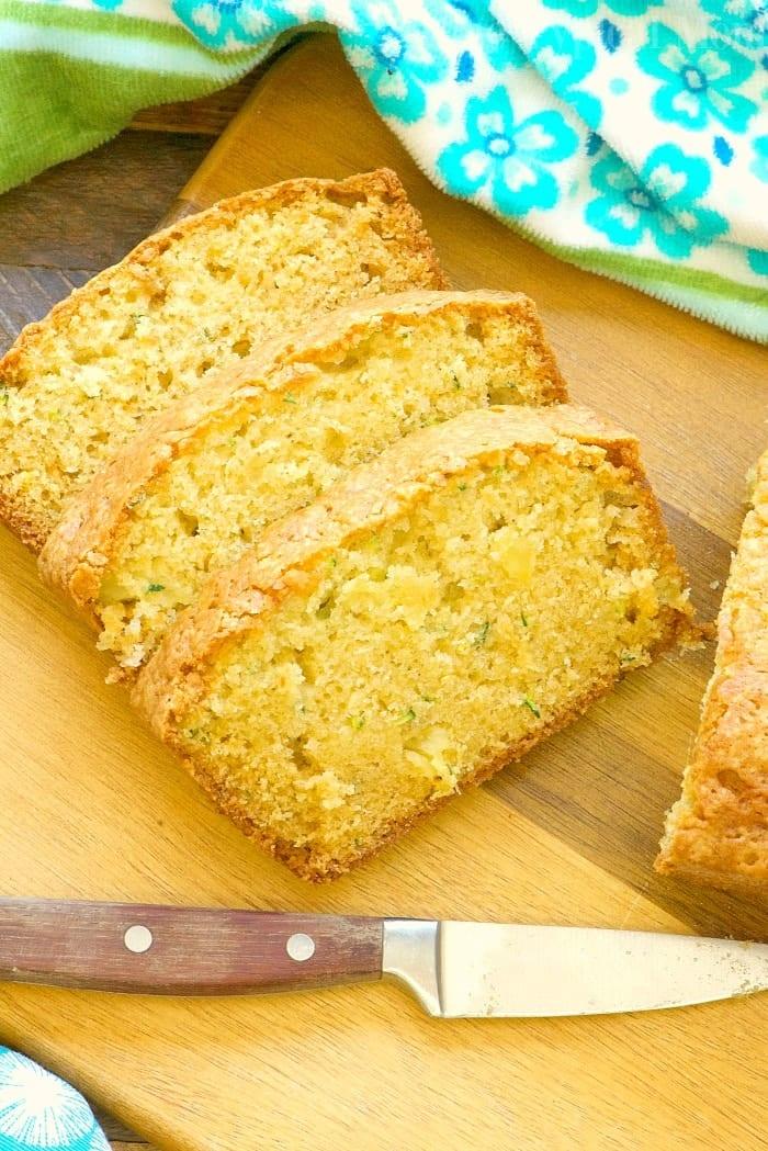 Sliced zucchini pineapple bread on a wooden cutting board next to a knife with a dark handle. A green and blue floral towel is partially visible in the background.