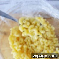 Effortless Microwave Pasta 8 A fork lifts cooked macaroni from a glass bowl covered with plastic wrap on a wooden surface, embodying the simplicity of how to cook pasta in the microwave.