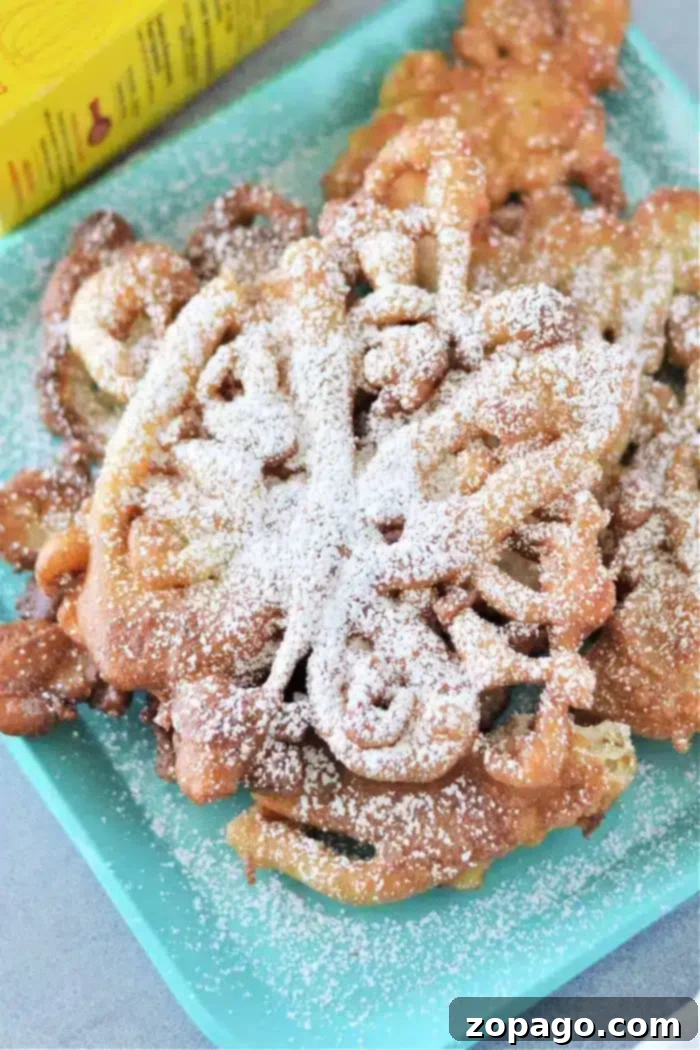 Effortless 3-Ingredient Bisquick Funnel Cake 2 A plate of golden-brown Bisquick funnel cakes, beautifully dusted with powdered sugar, rests on a teal square dish. The intricate swirling patterns of the cakes are visible, suggesting a perfect crispy texture. A yellow Bisquick box is partially visible in the soft-focus background, hinting at the key ingredient.
