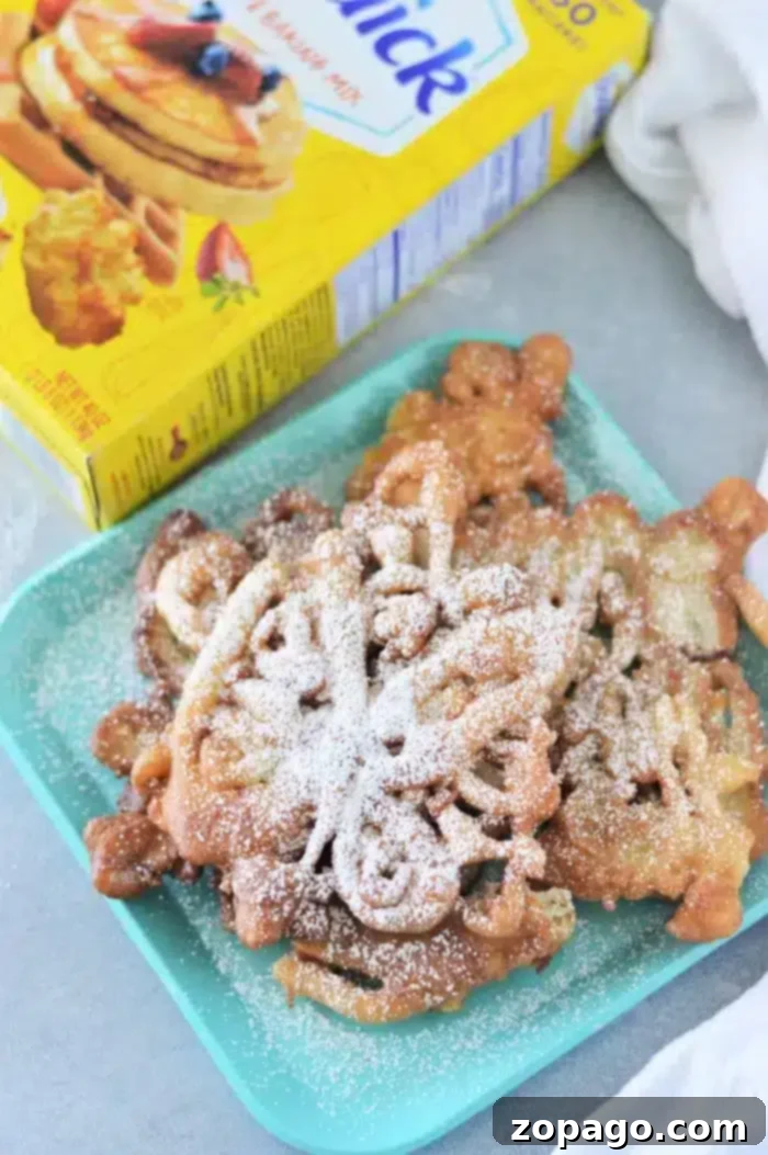 Effortless 3-Ingredient Bisquick Funnel Cake 6 A plate of golden-brown Bisquick funnel cakes is elegantly dusted with a generous amount of powdered sugar, with a yellow box of pancake mix resting nearby. The scene suggests a fresh, homemade treat ready for enjoyment.