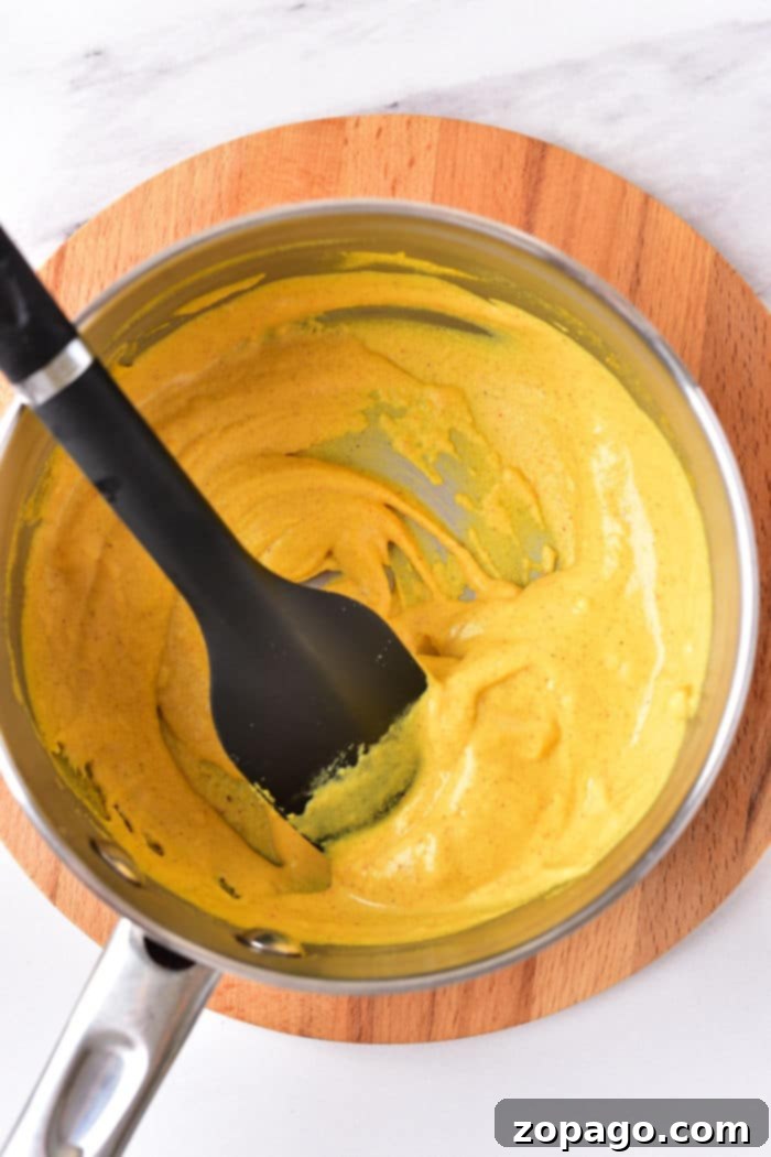 Crafting Your Perfect Hot Dog Mustard 5 A saucepan filled with creamy hot dog mustard, being stirred by a black spatula on a wooden trivet, demonstrating the cooking process.