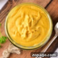 Glass jar of creamy yellow hot dog mustard on a wooden board with a spoon beside it.