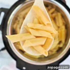 One-Pot Pressure Cooker Pasta 6 A wooden spoon holds perfectly cooked penne pasta, lifted gracefully above a pot. This delightful sight brings to mind the convenience and efficiency of preparing meals like pressure cooker pasta, where flavors meld seamlessly and every bite promises savory satisfaction.