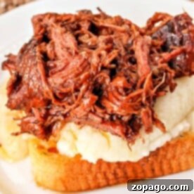 Ultimate Beef Manhattan 7 A savory Beef Manhattan graces the table, featuring a slice of toasted bread topped with creamy mashed potatoes and shredded beef, all elegantly presented on a white plate with a woven placemat underneath.