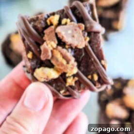 Close-up of a hand holding a decadent chocolate fudge, drizzled with rich chocolate and topped with crushed candy. The fudgy texture, enhanced by hints of peanut butter and creamy condensed milk, makes it a delightful indulgence.