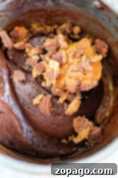 A thick chocolate fudge batter in a bowl is enhanced with chopped peanut butter cups and a dollop of creamy peanut butter, reminiscent of a rich fudge infused with the sweetness of condensed milk.