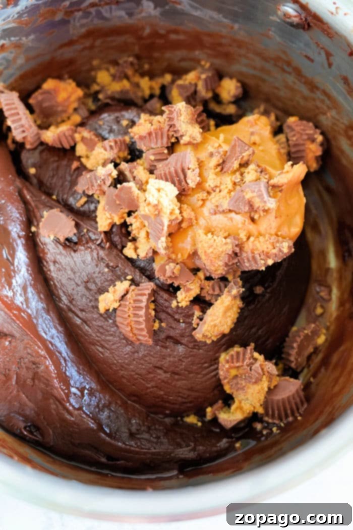 Decadent Peanut Butter Chocolate Fudge 9 A thick chocolate fudge batter in a bowl is enhanced with chopped peanut butter cups and a dollop of creamy peanut butter, reminiscent of a rich fudge infused with the sweetness of condensed milk.