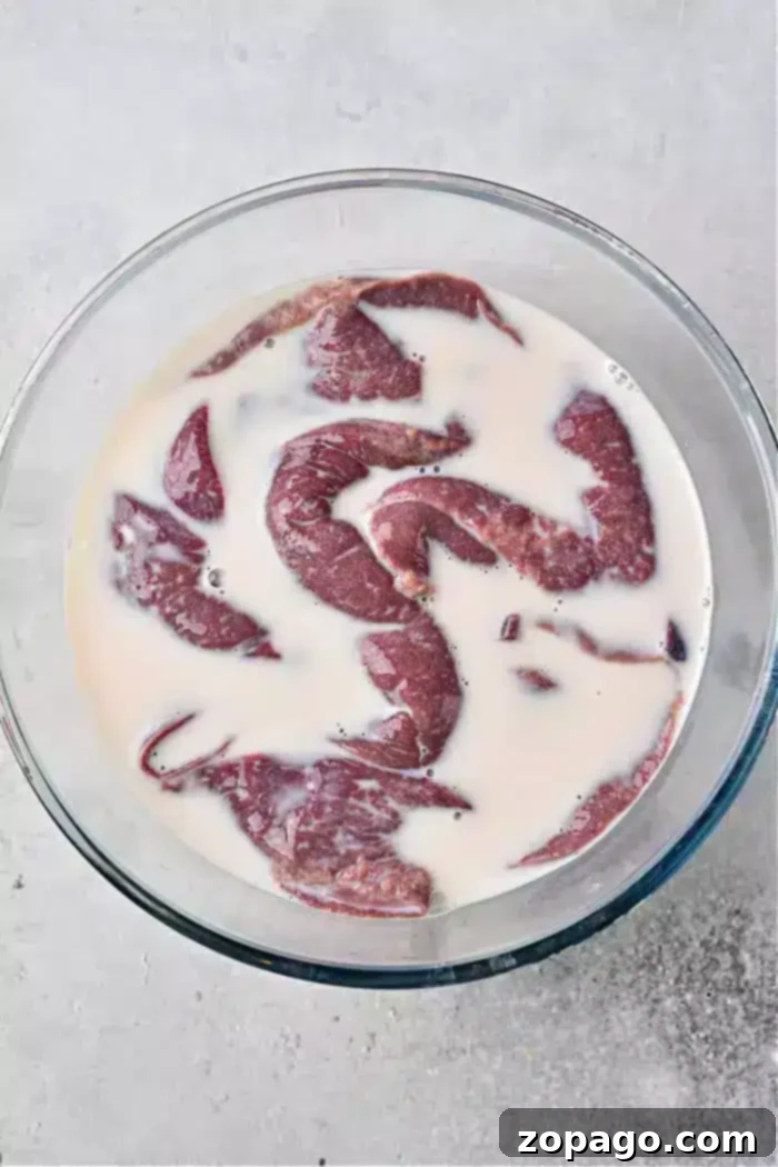 Delicious Pan-Seared Beef Liver 4 A glass bowl filled with pieces of raw beef liver soaking in milk rests on a light-colored, textured surface, essential for tenderizing.