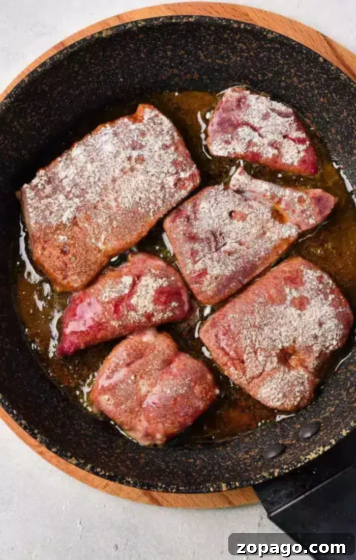Delicious Pan-Seared Beef Liver 5 A frying pan on the stove with six pieces of flour-coated beef liver cooking in hot oil, showcasing the ideal pan-frying process.
