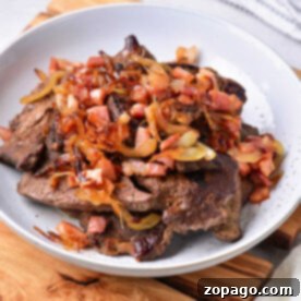 A white plate featuring a delicious dish of pan-fried beef liver topped with caramelized onions, diced tomatoes, and crispy bacon bits, served on a rustic wooden board.