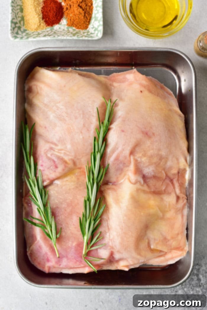 Juicy Baked Frozen Chicken Thighs 3 Raw frozen chicken thighs with rosemary sprigs rest in a tray, ready for baking. Nearby, spices and oil await their turn to enhance the flavor. Perfect for transforming frozen chicken thighs into a delicious meal.