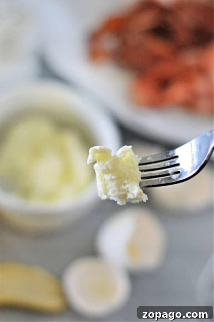 The Art of Perfect Egg Whites 6 A close-up of a fork holding a small piece of cooked egg whites. In the blurred background, a white bowl filled with more cheese sits beside a plate of sliced meat. Nearby, egg whites rest alongside garlic cloves, adding layers to the scene on the table.