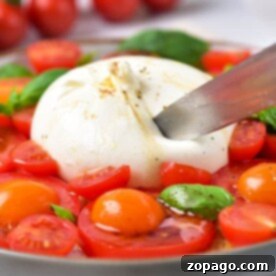 Burrata Caprese Nasıl Yapılır? 7 A knife slices through the creamy burrata in this Burrata Caprese dish, surrounded by juicy cherry tomatoes and fresh basil on a plate.