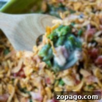 A wooden spoon lifts a serving of easy green bean casserole with crispy onions and diced ham from a green baking dish.
