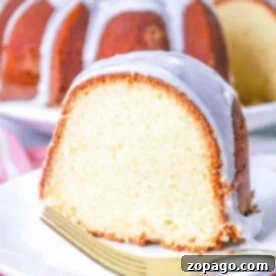 A perfectly glazed slice of bundt cake, a homemade version of Sara Lee Pound Cake, sits on a white plate with a gold fork, a pink floral cloth underneath.