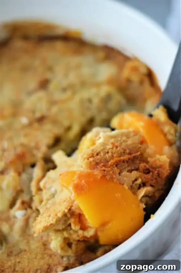Next-Level Box Cakes Secret Tips for a Homemade Taste 7 A close-up of a peach cobbler in a white baking dish, enhanced with clever box cake mix hacks, features a serving spoon lifting a portion. The cobbler boasts a golden-brown crust and visible peach slices.