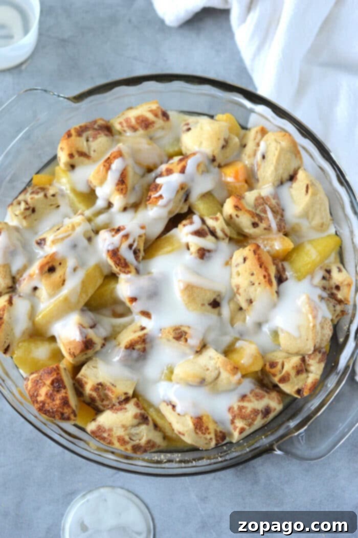Cinnamon Swirl Peach Cobbler 7 A glass dish filled with cinnamon roll biscuit pieces, peaches, and a drizzle of white icing evokes the comforting flavors of a cinnamon roll peach cobbler on a light gray surface.