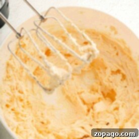 Easy Brownie Frosting 6 A mixing bowl filled with creamy, light yellow butter awaits. An electric mixer with two beaters, coated in the same velvety mixture for a luscious brownie frosting, rests above the bowl on a pristine white countertop.