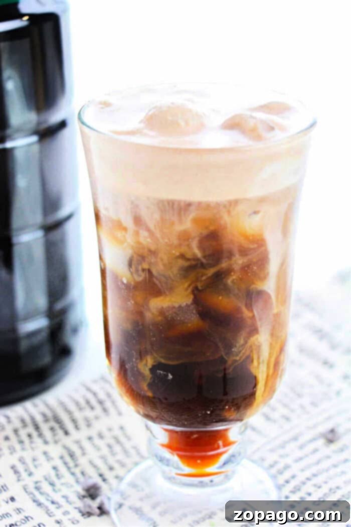 Whip Up Your Own Cold Foam 6 A glass filled with iced coffee topped with frothy cream sits on a surface covered with a newspaper, illustrating how to make cold foam for coffee. The drink has a layered appearance, showing dark coffee at the bottom and swirling milk throughout. A black bottle is partially visible in the background.