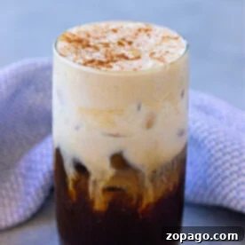 Whip Up Your Own Cold Foam 8 A glass of iced coffee with a creamy top, expertly frothed using a hand frother, and a sprinkle of cinnamon rests on a gray surface with a blue cloth nearby.