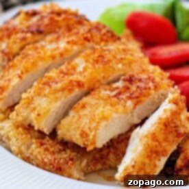 Parmesan-crusted chicken cutlet, perfectly cooked in the air fryer, sits sliced on a white plate, with fresh lettuce and tomato adding color to the background.