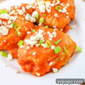 Three juicy buffalo chicken thighs topped with crumbled cheese and sliced green onions are artfully arranged on a pristine white plate.