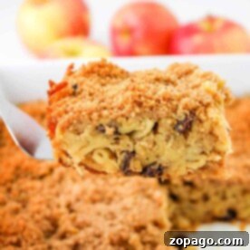A slice of apple kugel with a crumbly top is lifted from the baking dish, its golden crust inviting and perfectly textured. In the background, fresh apples add a vibrant touch, hinting at the delicious flavors within this classic treat.