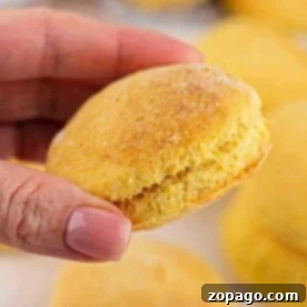 A hand holds a freshly baked golden cornmeal biscuit, with more biscuits blurred in the background.