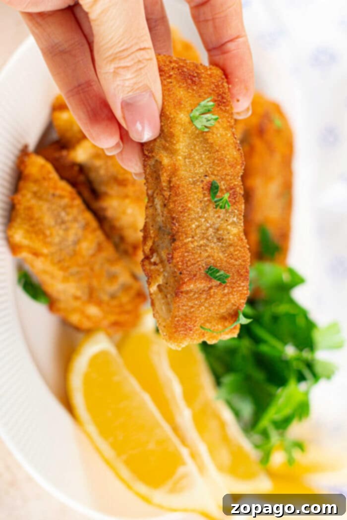 Cornmeal: The Key to Crispy Fish Fry 2 A hand holds a crispy fried piece of fish, perfectly coated in cornmeal for that extra crunch, over a plate adorned with lemon wedges and fresh greens.