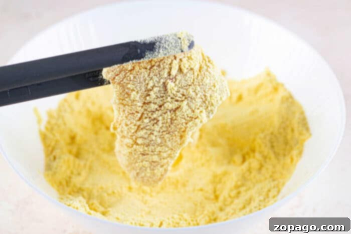 Cornmeal: The Key to Crispy Fish Fry 4 Black tongs hold a piece of food coated in seasoned yellow cornmeal for a fish fry, above a bowl filled with the same cornmeal mixture.