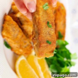 A hand holds a crispy fried piece of fish, perfectly coated in cornmeal for that extra crunch, over a plate adorned with lemon wedges and fresh greens.