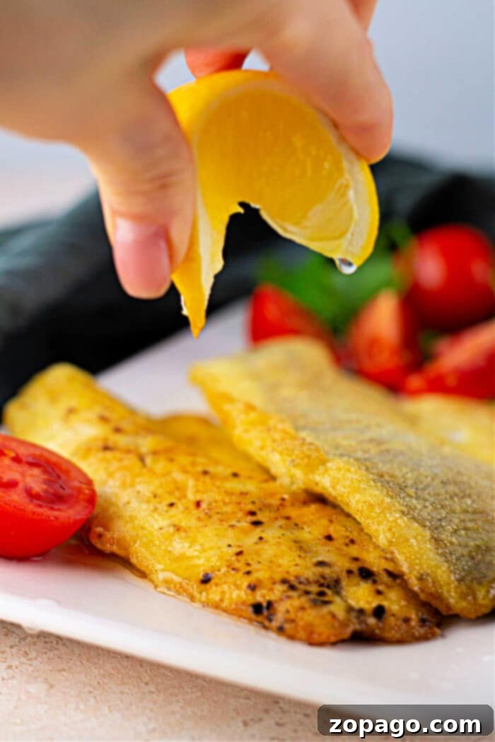 Golden Pan Fried Swai 6 Hand squeezing lemon over seasoned fried Swai fish with cherry tomatoes on a white plate.