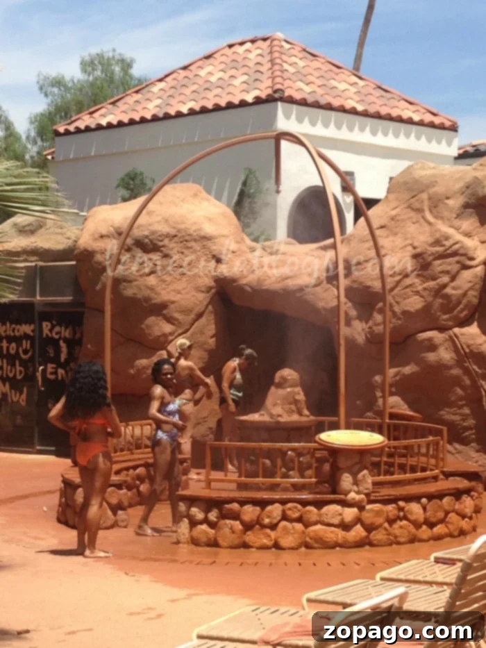 The best spas in Southern California! From Orange County, to San Diego, Los Angeles and everywhere in between you've got to go. People in swimsuits stand around a mud bath area with rock formations and a wooden structure. In the background, there is a building with a terracotta-tiled roof. A sign reading "Welcome to Club Mud" is visible. A few lounge chairs are in the foreground, showcasing one of the best spas in Southern California.