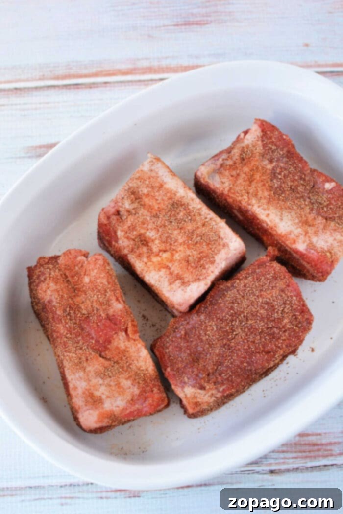 Perfect Oven Baked Beef Ribs 6 Four seasoned beef ribs in a white dish on a light wooden surface, ready to be compared to the tenderness you achieve with beef ribs in the oven at 275.