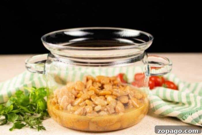From Dry to Delicious Butter Beans 5 A glass pot filled with butter beans sits beside fresh parsley and cherry tomatoes on a striped cloth, offering inspiration on how to cook butter beans with a touch of garden-fresh simplicity.