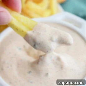 A hand dipping a fry into creamy, seasoned Blackened Ranch Sauce from Popeyes in a white bowl, with more fries in the background.