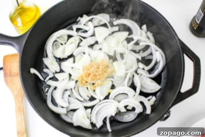 Perfectly Cooked Bratwurst Every Time 4 A cast iron skillet on a stovetop containing sliced onions and minced garlic, with steam rising from the pan. A wooden spatula is positioned next to the skillet, and a bottle of olive oil is visible in the background, setting the perfect stage for how to cook bratwurst to perfection.