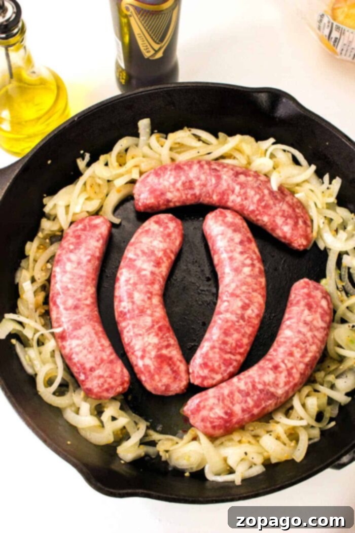 Perfectly Cooked Bratwurst Every Time 5 Five raw sausages are placed on a bed of sliced onions sizzling in a cast iron skillet, demonstrating how to cook bratwurst to perfection. A bottle of olive oil and a bottle of Guinness beer are visible in the background.