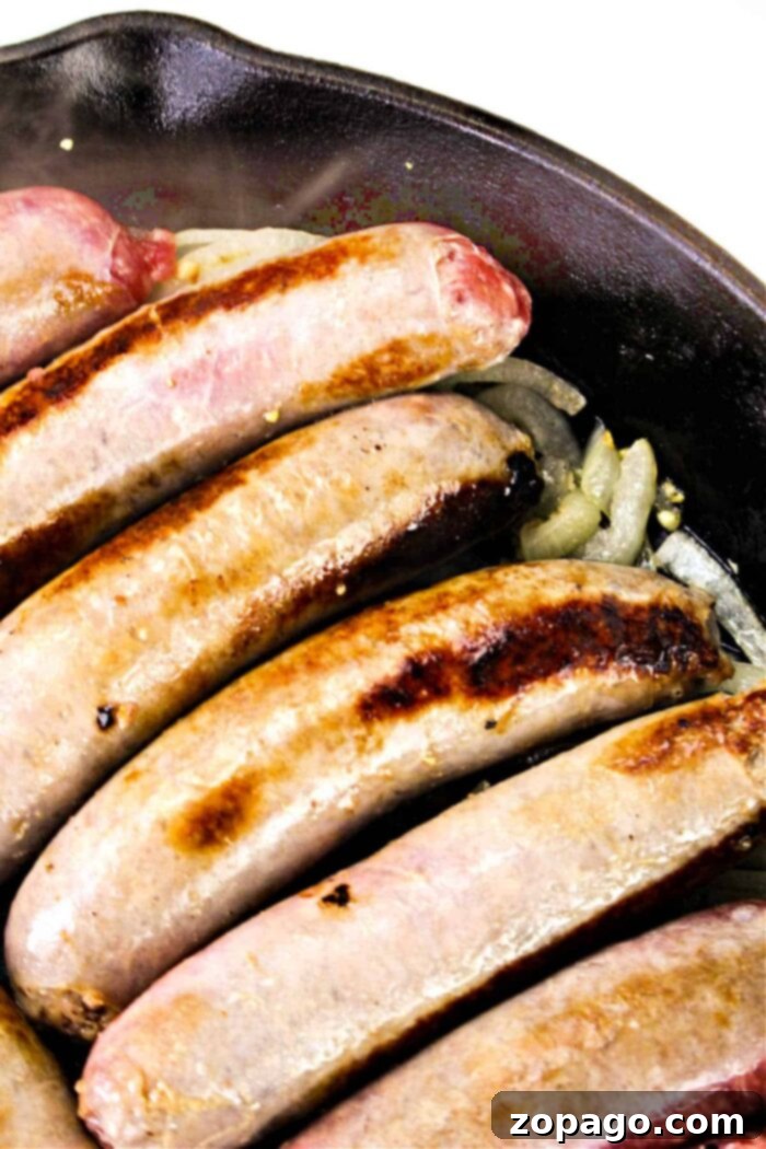 Perfectly Cooked Bratwurst Every Time 6 Five sausages are cooking in a cast iron skillet. The sausages have a browned, crispy exterior and are lying on a bed of sliced onions. The image shows the pan from an overhead perspective, offering a delectable hint on how to cook bratwurst to perfection.
