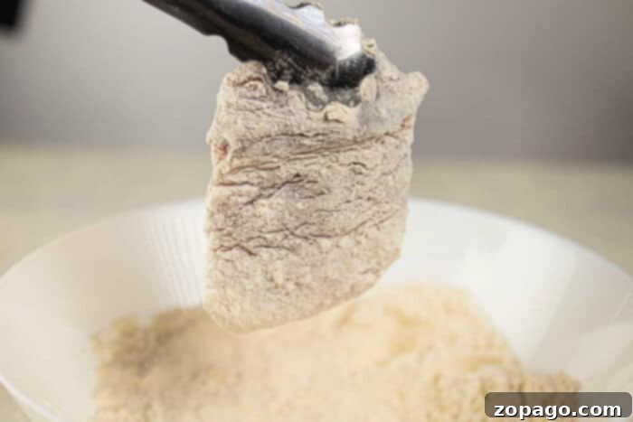 Golden Crispy Pork Chops 4 A piece of breaded boneless pork chops, soon to become a crispy delight, is held by tongs over a bowl of flour, ready to transform into savory deep-fried pork chops.